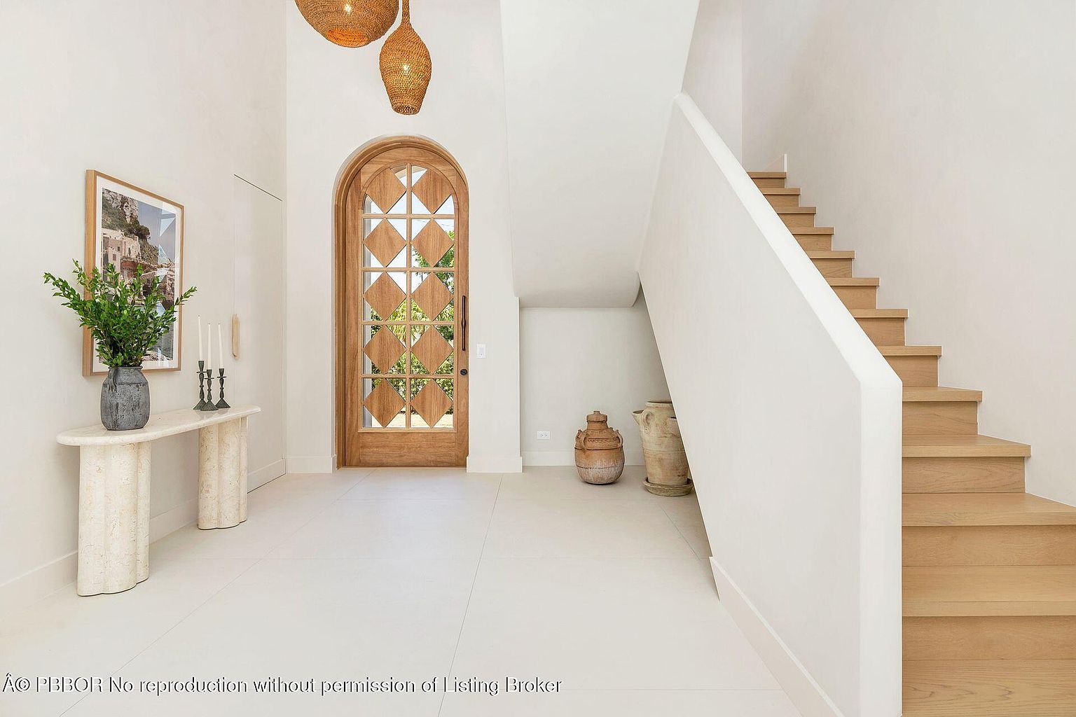 This is an interior shot of a bright and airy hallway featuring a unique arched wooden door with diamond-shaped glass panes. A travertine console table with a plant and candles sits to the left, while a staircase with light wood treads and a white banister ascends to the right. The white tile flooring and walls contribute to a clean, modern aesthetic.
