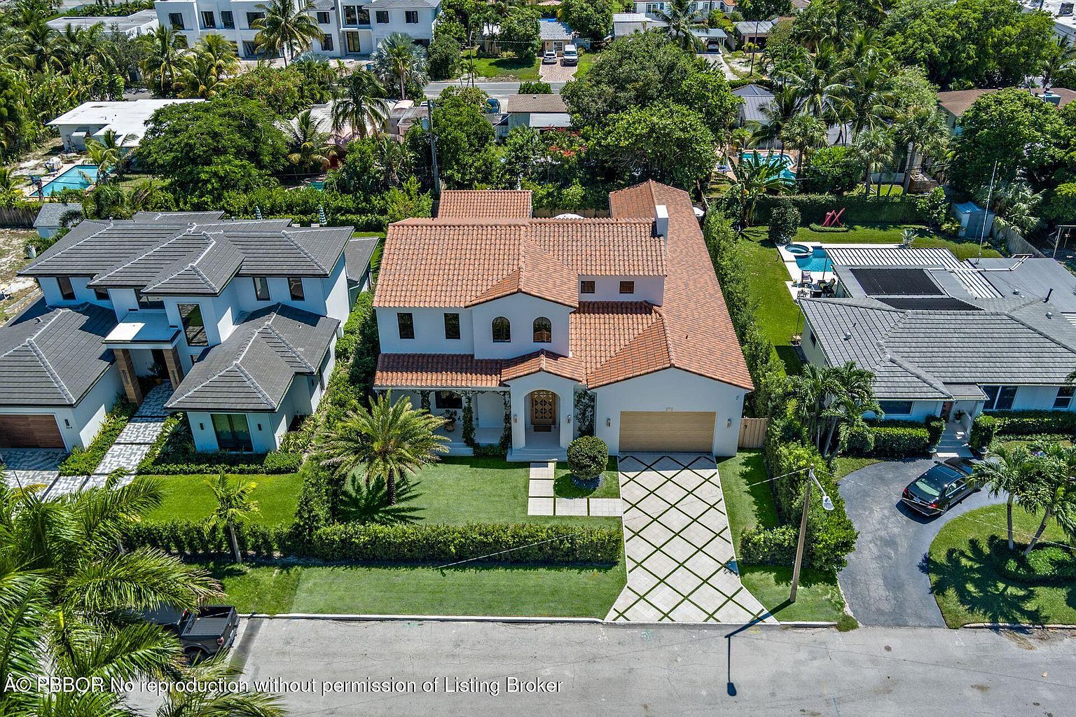 This aerial shot showcases a beautifully landscaped property featuring a Mediterranean-style home with a terracotta tile roof. The meticulously maintained lawn, patterned driveway, and mature trees enhance the curb appeal. The surrounding neighborhood is lush and residential, creating a serene and upscale atmosphere.