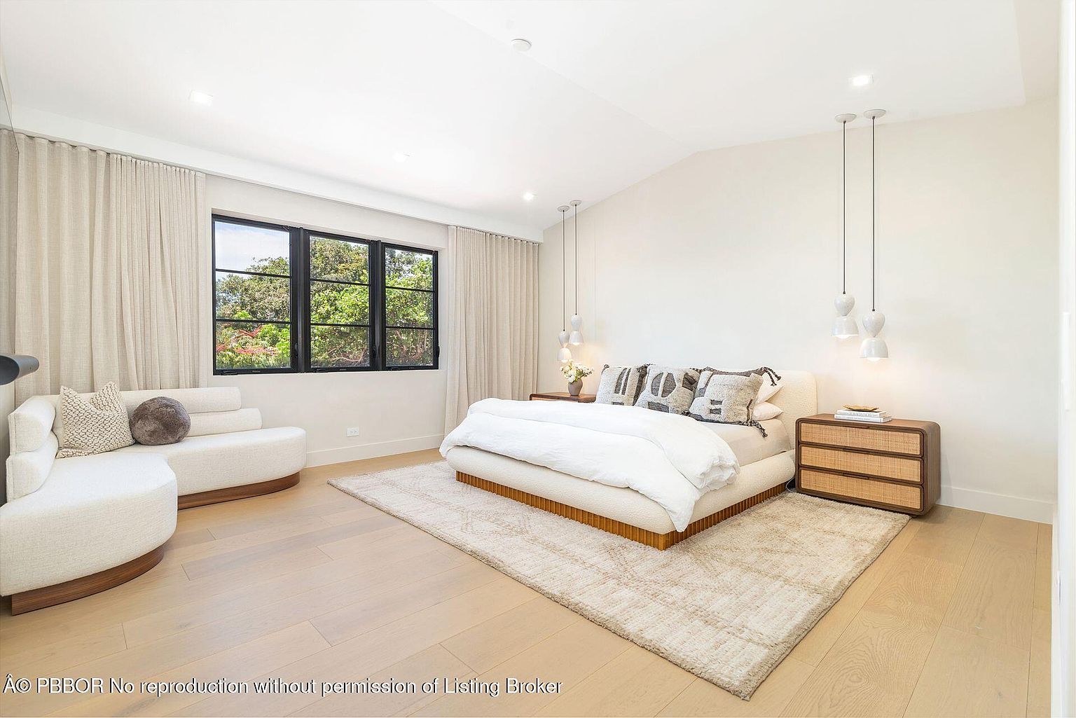 This is a bright and airy primary bedroom featuring a large bed with neutral-toned bedding and decorative pillows. The room is well-lit with natural light from a window and complemented by modern pendant lighting. A curved sofa and a wooden nightstand add to the room's sophisticated and comfortable ambiance.