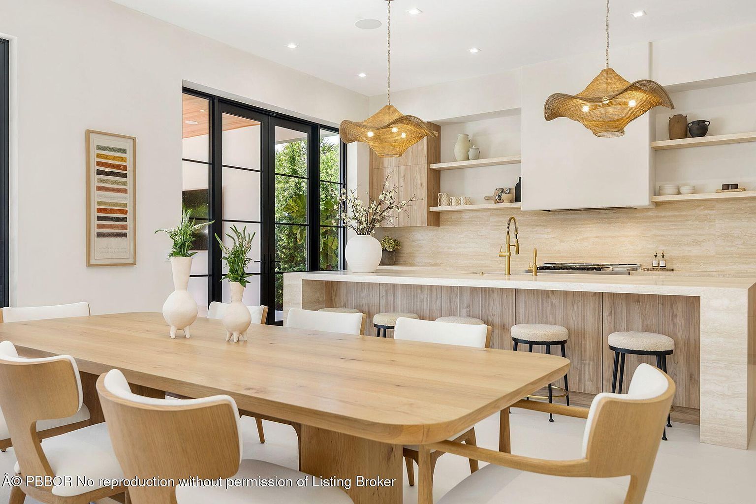 This interior shot showcases a dining area with a large wooden table surrounded by modern chairs with white cushions. Two decorative vases adorn the table, and unique pendant lights hang above. The space is bright and airy, with a view of greenery through the black-framed windows, creating an inviting and stylish dining experience.