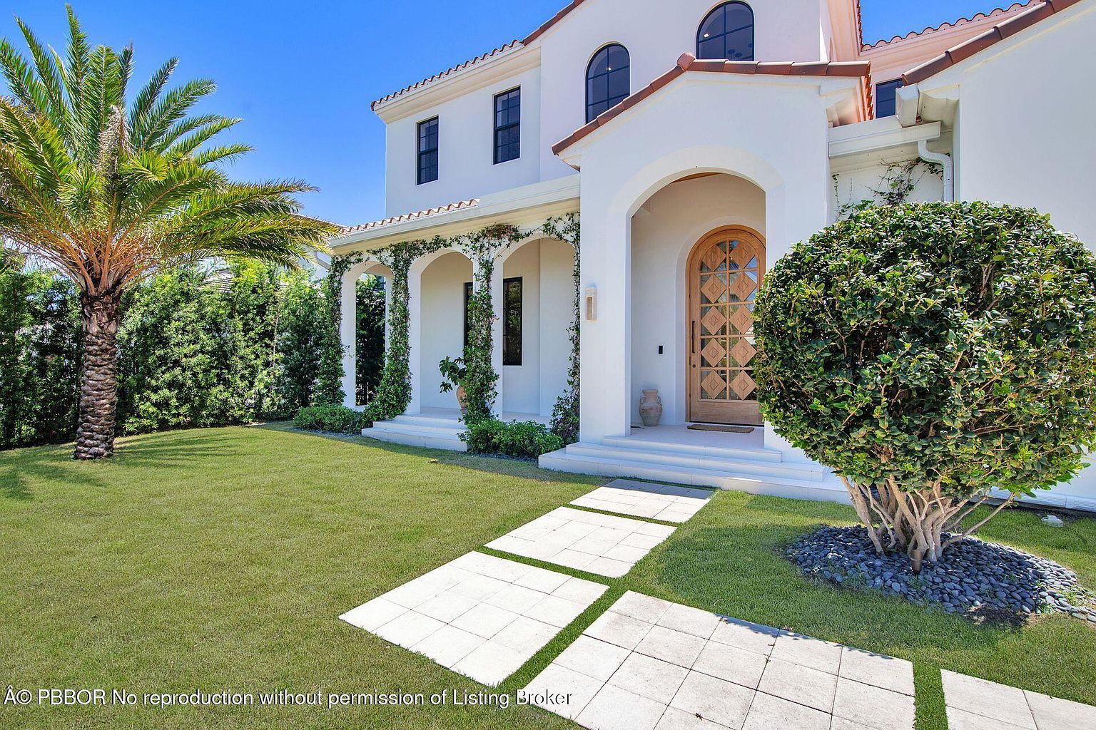 This is a front exterior view of a luxurious two-story home with a Mediterranean architectural style. The house features a white stucco facade, a red tile roof, arched windows, and a covered entryway with climbing vines on the columns. A well-manicured lawn, a palm tree, and a stone pathway leading to the front door enhance the property's curb appeal.