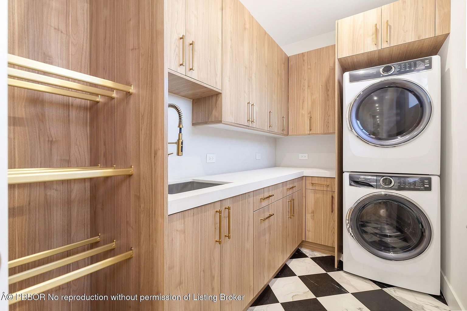 This is a well-organized laundry room featuring light wood cabinetry with gold hardware, a white countertop, and a stainless steel sink with a gold faucet. A stacked washer and dryer unit is positioned against the wall, and a clothing rack is built into the cabinetry on the left. The black and white checkered floor adds a touch of classic style.