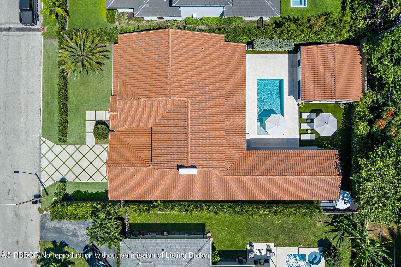 This aerial shot showcases a luxurious property with a terracotta-tiled roof, a sparkling blue swimming pool, and meticulously landscaped grounds. The house features a U-shaped design, complemented by a detached structure near the pool area. The driveway is paved with a geometric pattern, adding to the property's curb appeal.