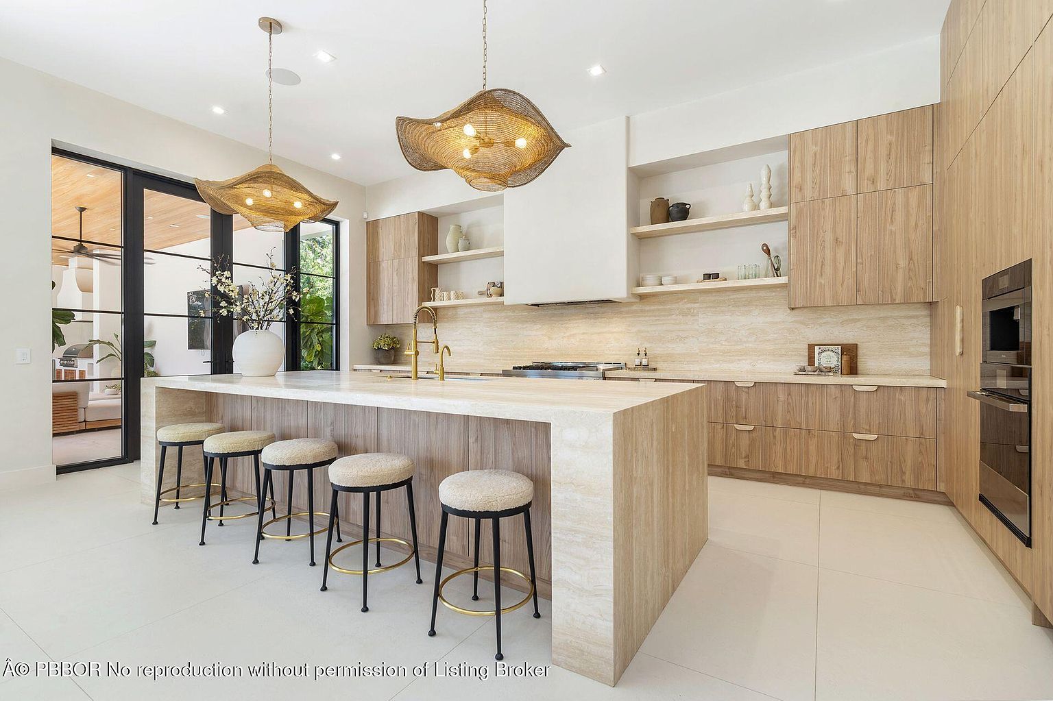 This is a bright and modern kitchen featuring light wood cabinetry and a large island with a stone countertop and seating. The kitchen is illuminated by unique pendant lighting and natural light from a large window. The overall impression is clean, spacious, and luxurious, highlighting the high-end finishes and contemporary design.