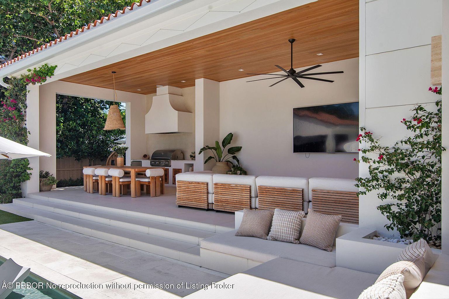 This image showcases a luxurious outdoor living space featuring a covered patio with a wooden ceiling and a built-in outdoor kitchen. A dining area with a wooden table and chairs is visible, along with a comfortable seating area with plush cushions and throw pillows. The space is well-lit and features a large television, creating an inviting atmosphere for relaxation and entertainment.