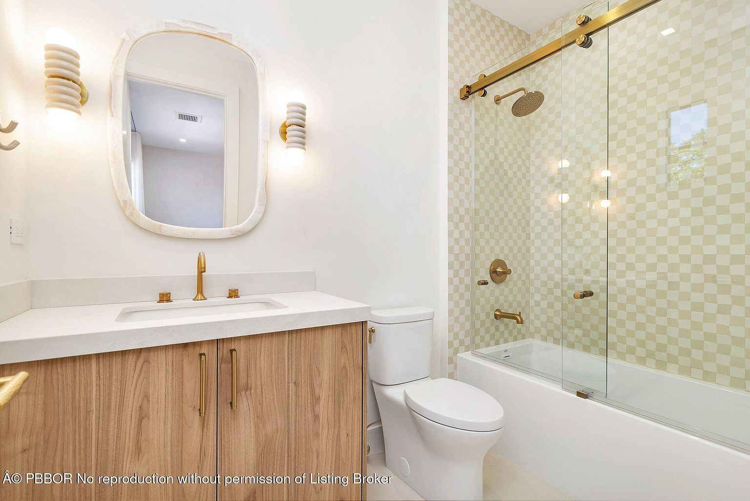 This is a well-lit bathroom featuring a modern design. The vanity has a light wood cabinet with gold hardware and a white countertop with an integrated sink. A unique mirror hangs above the vanity, flanked by sconce lighting. The bathroom also includes a toilet and a shower/tub combination with a glass door and gold fixtures, all set against a patterned tile wall.