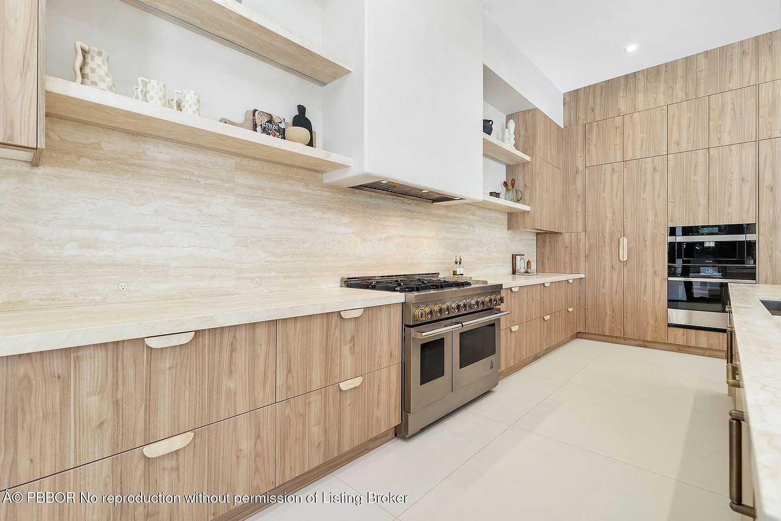 This is a modern kitchen featuring light wood cabinetry and drawers with unique handle designs. A stainless steel range is the focal point, complemented by a white range hood and open shelving. The countertops and backsplash are a light, textured stone, and the floor is tiled in a light color, creating a bright and airy atmosphere.