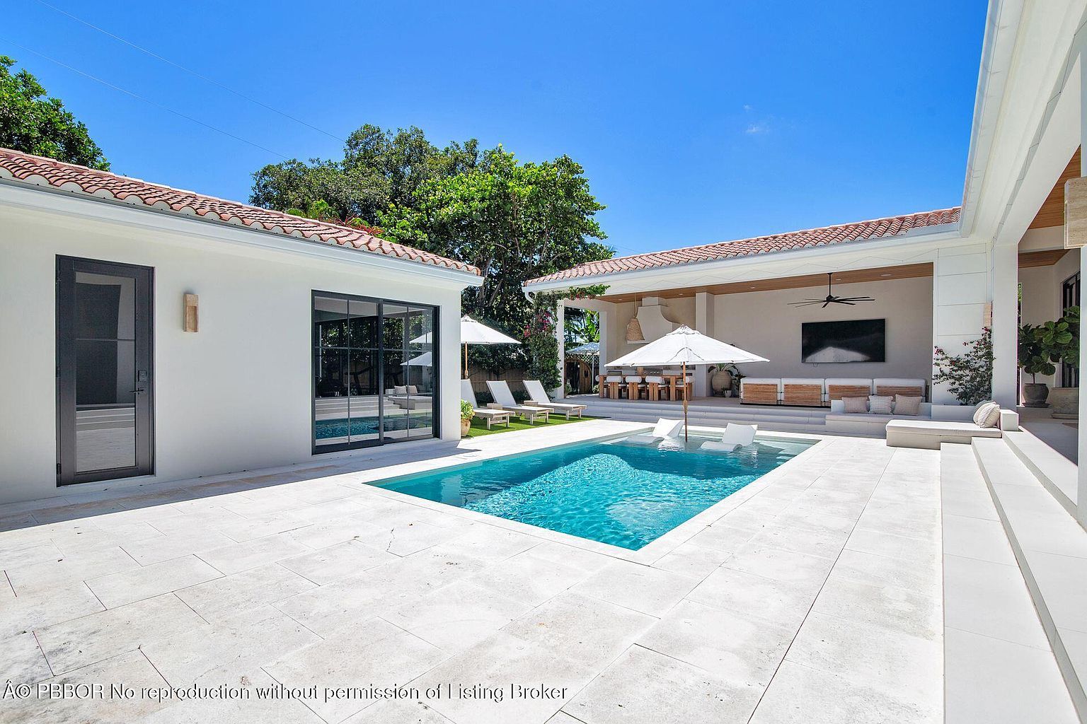 This image showcases a luxurious backyard pool area, featuring a rectangular pool with clear blue water, surrounded by light-colored stone tiling. The pool is flanked by modern white buildings with terracotta roofs, one of which includes a covered outdoor living space with a large television and comfortable seating. The scene is bathed in sunlight, creating an inviting and upscale ambiance.