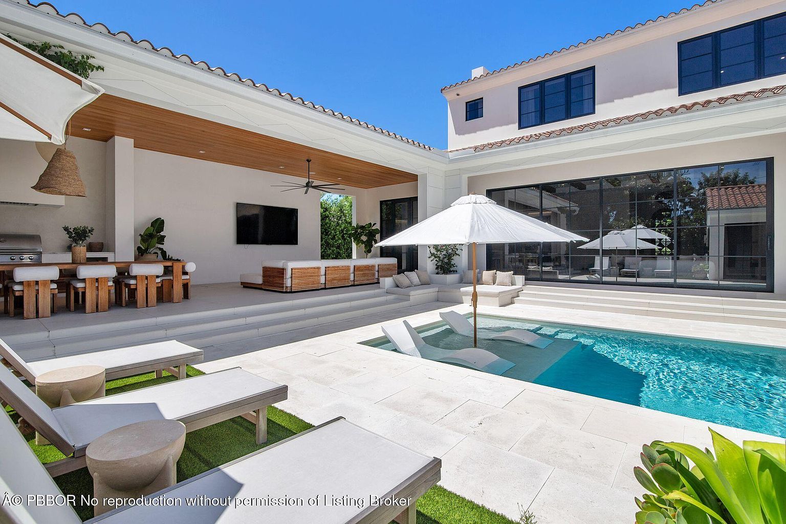 This image showcases a luxurious outdoor pool and patio area. The pool features built-in lounge chairs and is surrounded by a pristine white stone patio. The patio includes a covered dining area with a grill, a seating area with a large television, and additional lounge chairs, creating an inviting and upscale outdoor living space.