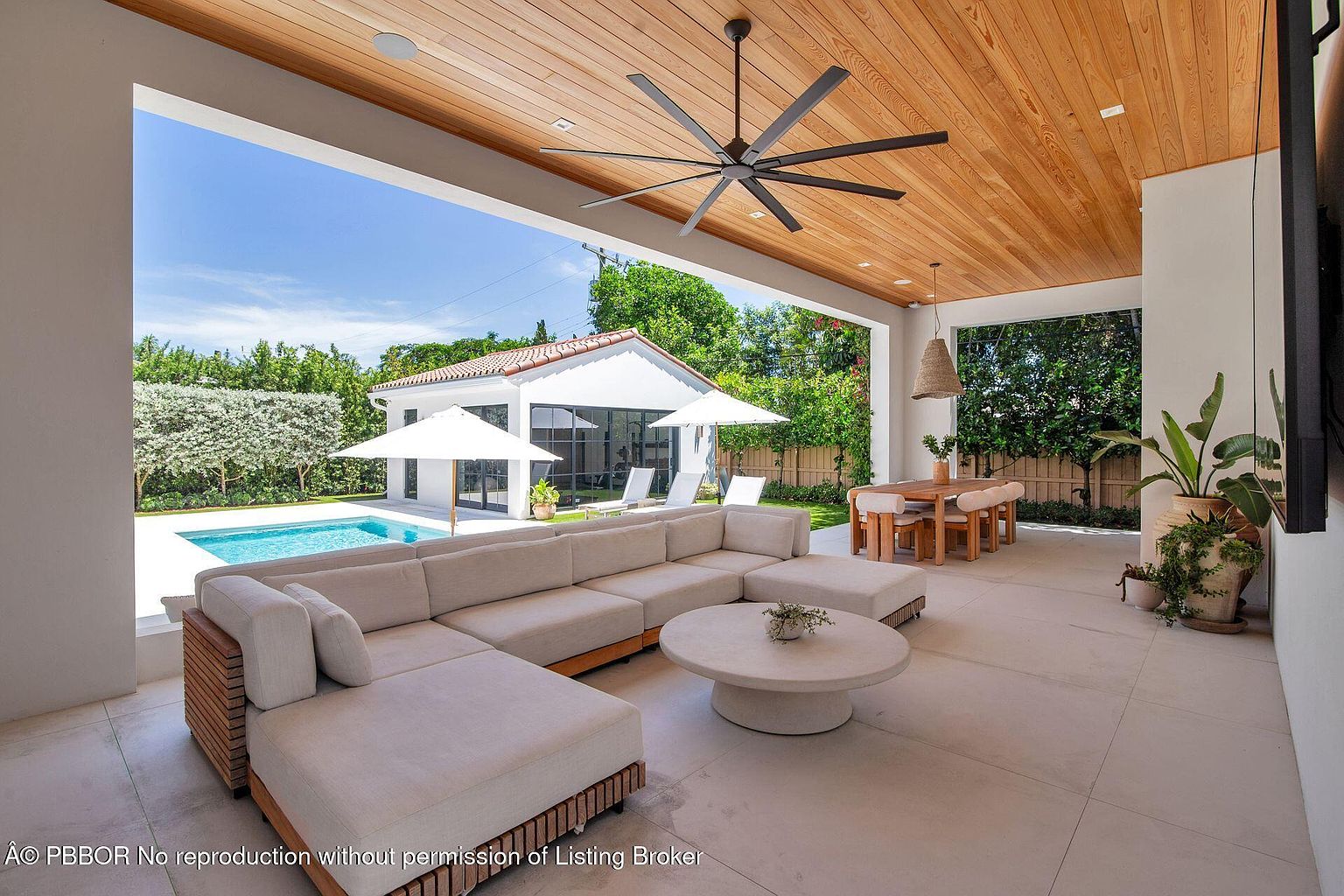 This image showcases a luxurious outdoor patio area, featuring a large sectional sofa and a round coffee table under a wooden ceiling with a ceiling fan. The patio overlooks a pool and a well-manicured garden, with a glimpse of a guest house in the background. The space is designed for relaxation and entertainment, blending indoor comfort with outdoor beauty.
