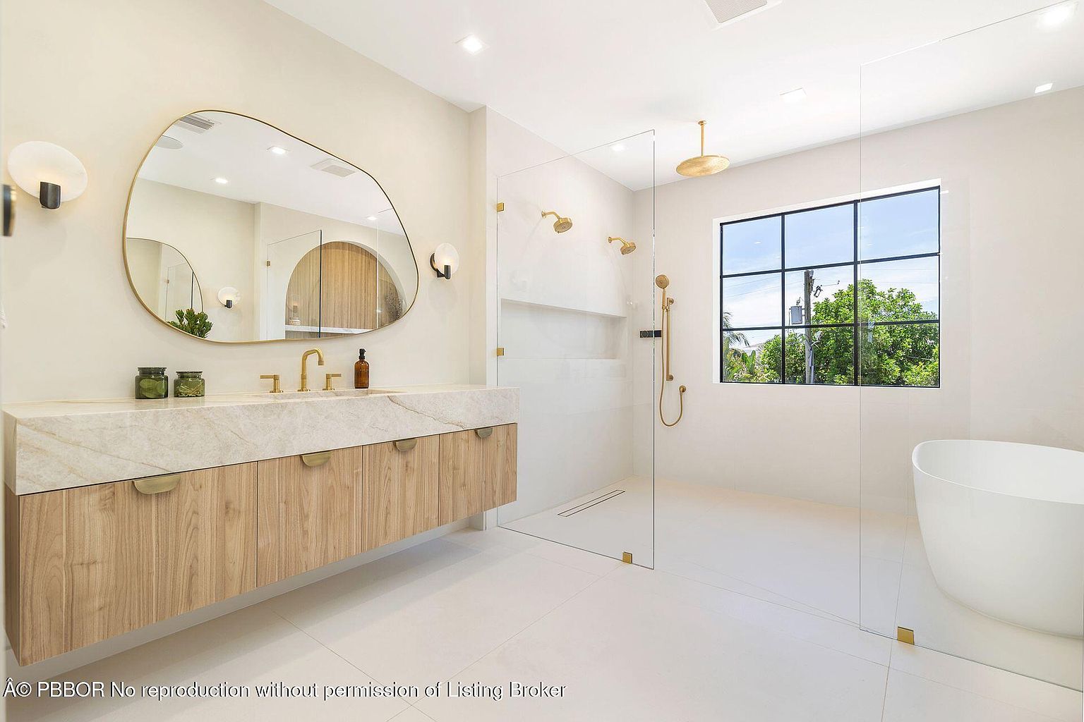 This is a bright and modern primary bathroom featuring a floating vanity with a light wood finish and a marble countertop. An organically shaped mirror hangs above the vanity, flanked by sconces. The bathroom also includes a glass-enclosed shower with gold fixtures and a freestanding bathtub, all set against a backdrop of white tile and walls, creating a clean and luxurious space.