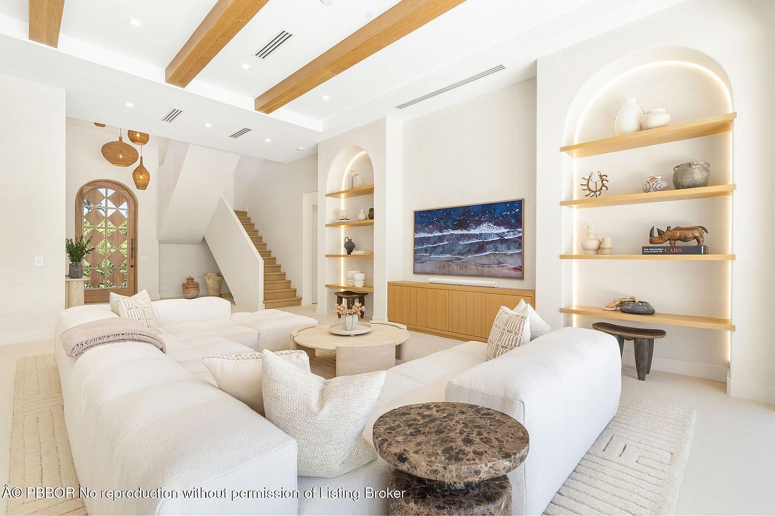 This is a bright and airy living room featuring a large, modular white sofa, a round coffee table, and built-in shelving with decorative items. The room has a modern, minimalist style with wooden ceiling beams and an arched doorway leading to a staircase. A large television is mounted above a wooden console, and the overall impression is one of comfort and sophistication.