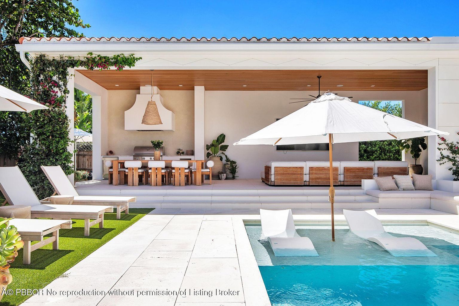 This image showcases a luxurious outdoor living space with a pool featuring in-water lounge chairs. The area includes a covered patio with a dining area, a built-in grill, and comfortable seating. The design emphasizes a seamless blend of indoor and outdoor living, perfect for relaxation and entertainment.