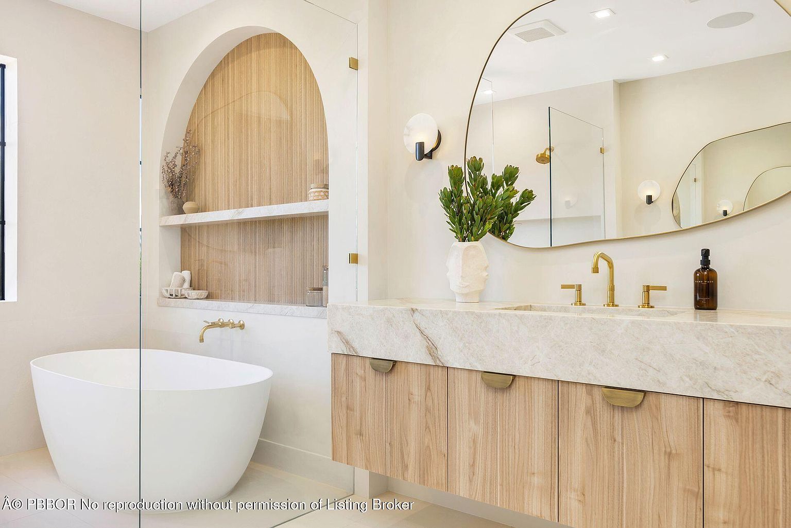 This is a luxurious primary bathroom featuring a freestanding white bathtub and a modern vanity with a marble countertop and light wood cabinetry. An arched alcove with shelving adds architectural interest, while a large, uniquely shaped mirror and stylish light fixtures enhance the contemporary design. The space is bright, clean, and designed for relaxation.