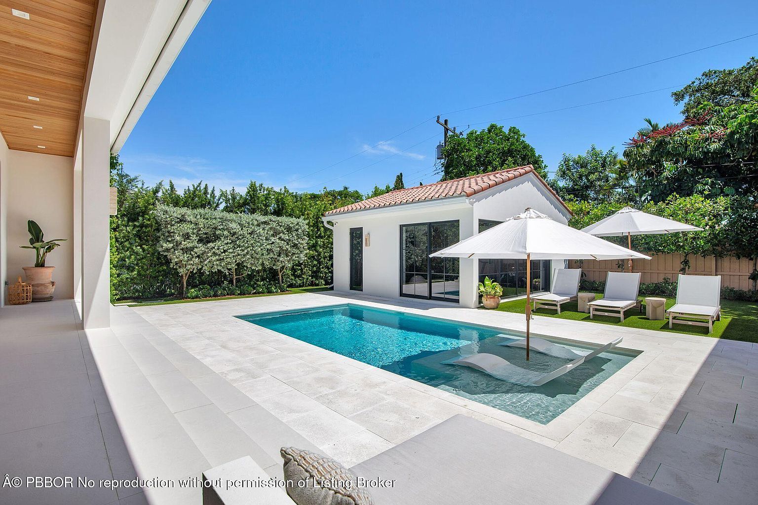 This image showcases a luxurious backyard pool area, featuring a rectangular pool with a shallow sunbathing area. The pool is surrounded by light-colored stone tiling and complemented by modern lounge chairs and white umbrellas. A white pool house with black-framed glass doors adds a touch of elegance, while lush greenery provides privacy and a serene backdrop.