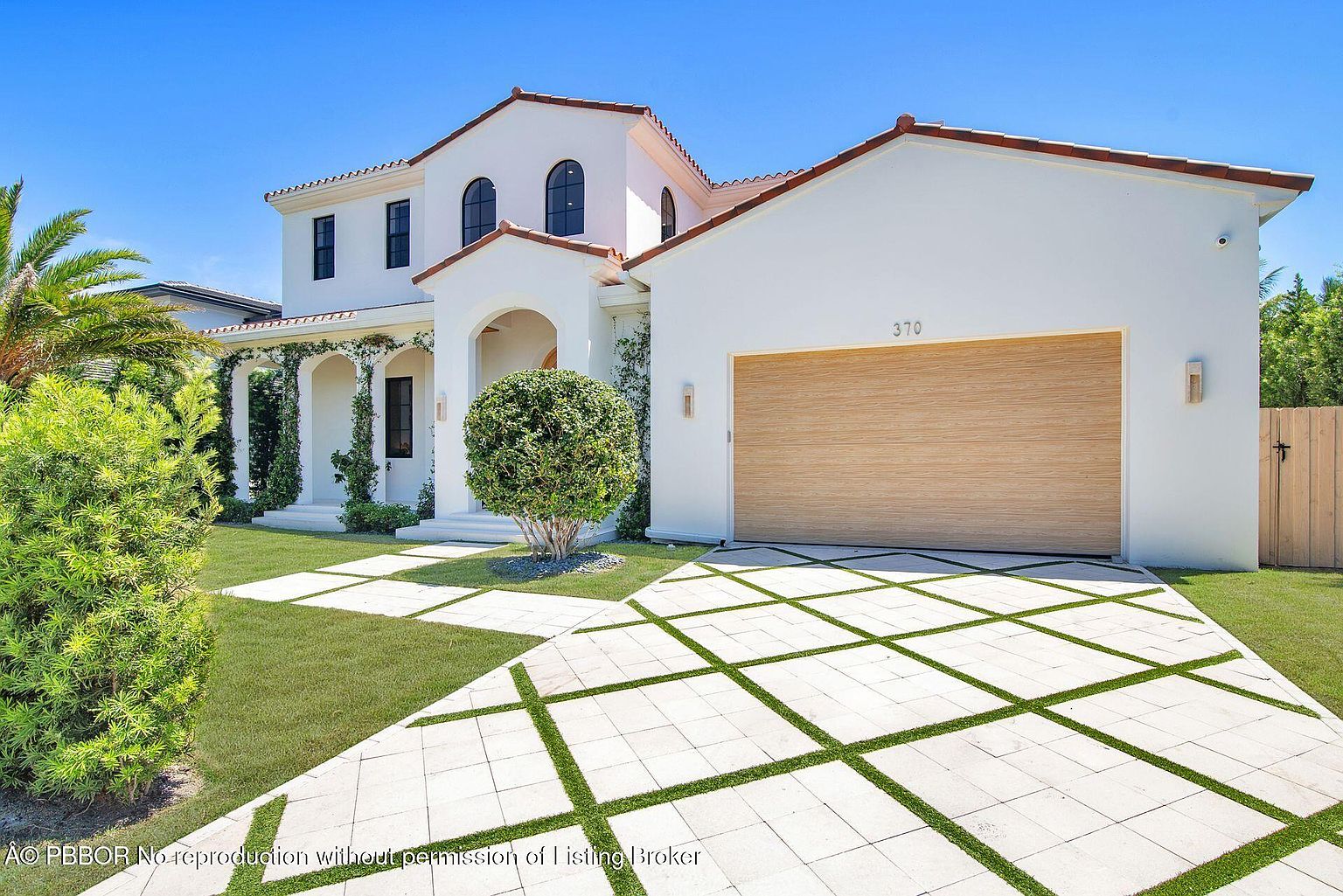 This is a front exterior view of a two-story Mediterranean-style home. The house features a white stucco exterior, a red tile roof, arched entryways, and a wooden garage door. The driveway is paved with light-colored stones interspersed with grass, and the landscaping includes manicured bushes and palm trees, creating a luxurious and inviting curb appeal.
