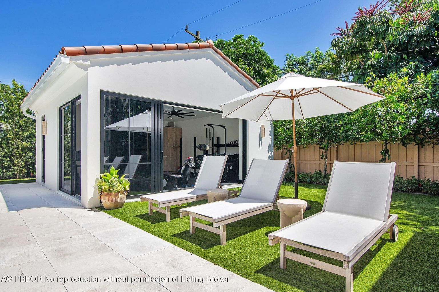 This image showcases a modern backyard featuring a white building with black-framed sliding glass doors, which appears to be a home gym. The outdoor space includes a well-manicured lawn, three lounge chairs with a small side table and a large umbrella, creating a relaxing and inviting atmosphere. The scene is framed by lush greenery and a wooden fence, enhancing the sense of privacy and tranquility.