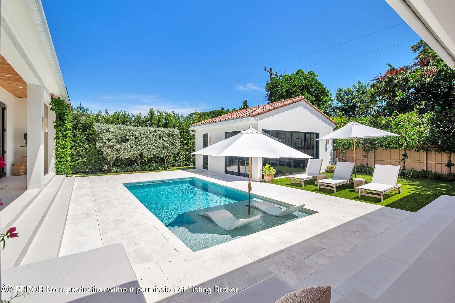 This image showcases a luxurious backyard pool area. The pool features a shallow lounging area with in-water loungers and is surrounded by white tile. The backyard also includes lounge chairs with umbrellas on a manicured lawn, complemented by a modern white building with a red tile roof, creating an inviting and upscale outdoor living space.