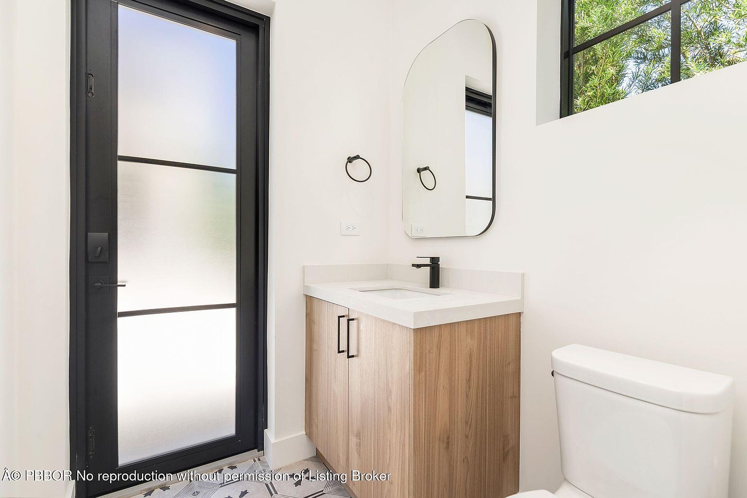 This is a modern guest bathroom featuring a wood-toned vanity with a white countertop and a black faucet. A sleek, arched mirror hangs above the sink, complemented by black towel rings. The room is painted in a clean white, and the floor is tiled with a geometric pattern, creating a stylish and inviting space.