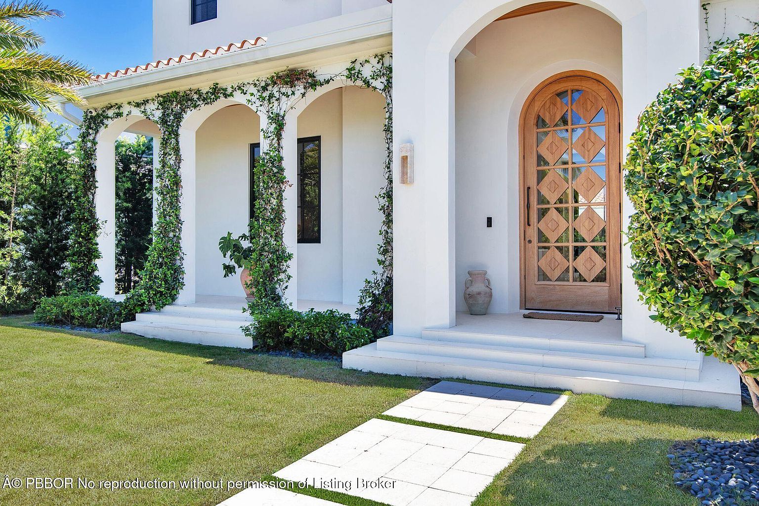 This image showcases the elegant entryway of a home, featuring a striking arched wooden door with diamond-patterned glass panels. The entrance is framed by white columns adorned with climbing greenery, leading to a well-manicured lawn and a stone pathway. The overall impression is one of sophisticated curb appeal and inviting warmth.