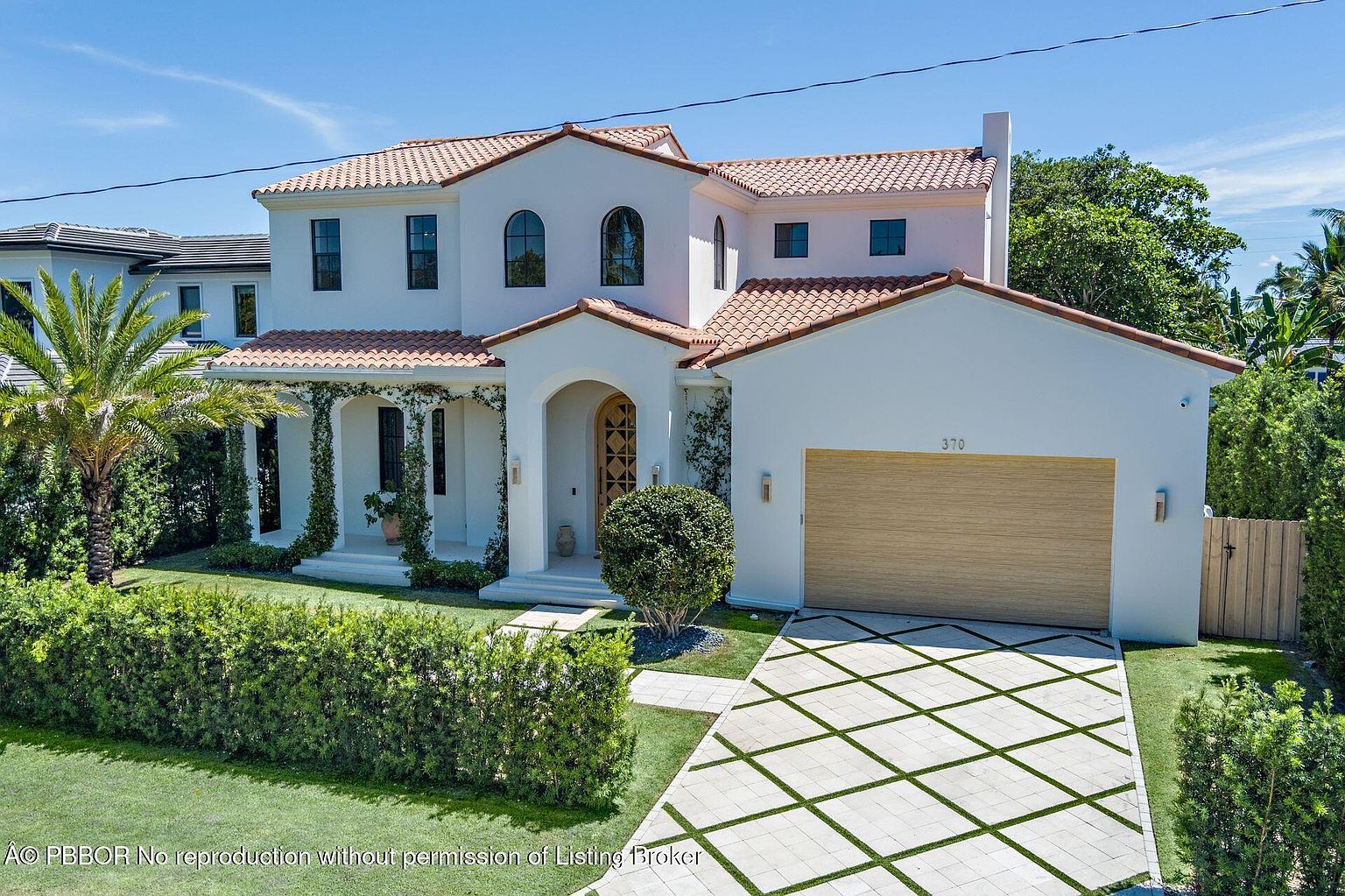 This is a front exterior view of a two-story Mediterranean-style home. The house features a red tile roof, white stucco walls, and arched windows. A well-manicured lawn, hedges, and a patterned driveway enhance the property's curb appeal, creating an inviting and luxurious impression.