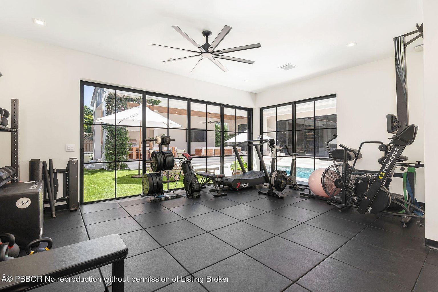 This is an interior shot of a home gym featuring black rubber flooring and large windows that offer a view of the backyard and pool area. The gym is equipped with various exercise machines, including a treadmill, rowing machine, and weightlifting equipment. The room is well-lit and appears spacious, creating an inviting atmosphere for workouts.