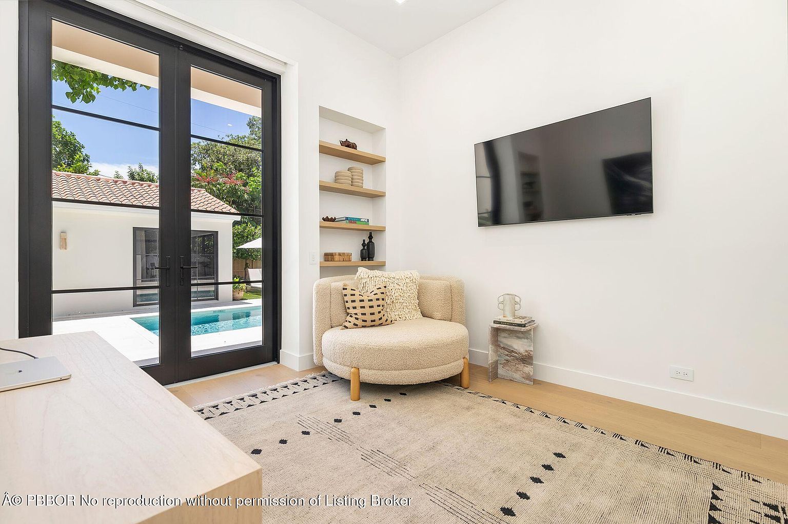 This interior shot showcases a modern office or study area with a focus on natural light and minimalist design. A black-framed glass door provides a view of the pool area, while a built-in shelving unit and a wall-mounted TV add functionality. The room is furnished with a round chair, a marble side table, and a patterned rug, creating a comfortable and stylish workspace.