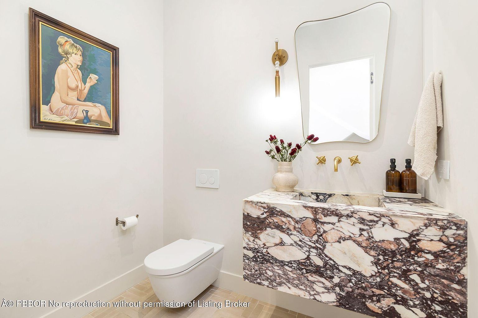 This is a stylish powder room featuring a unique marble vanity with a vessel sink and gold fixtures. A modern, asymmetrical mirror hangs above the sink, complemented by a sconce. The room also includes a wall-mounted toilet and a framed art piece, creating a sophisticated and contemporary space.