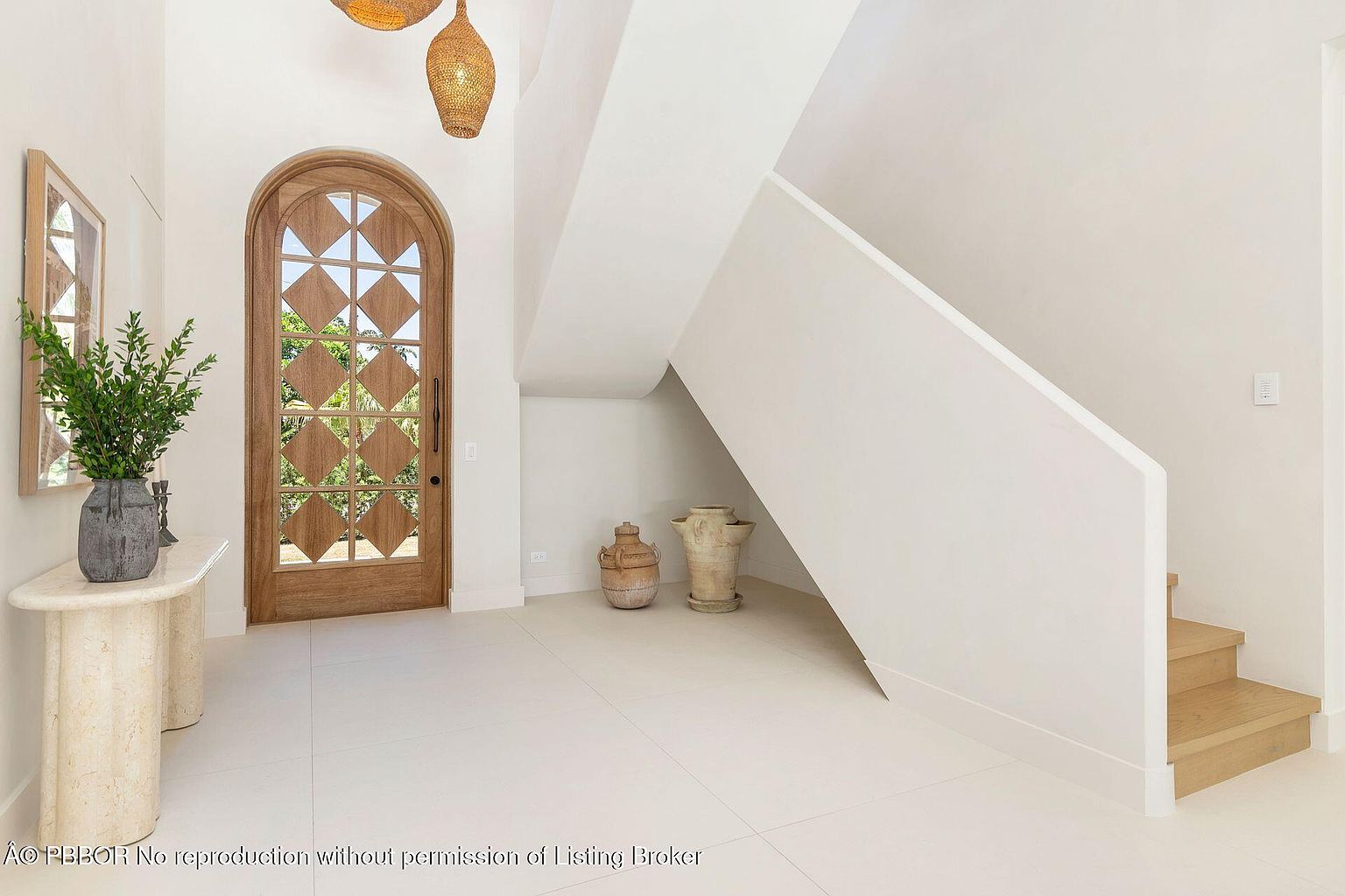 This interior shot showcases a bright and airy hallway with a striking arched wooden door featuring diamond-patterned glass. A travertine console table with a plant adds a touch of elegance, while the staircase with a minimalist design leads to the upper level. The white walls and light tile flooring contribute to the spacious and clean aesthetic.
