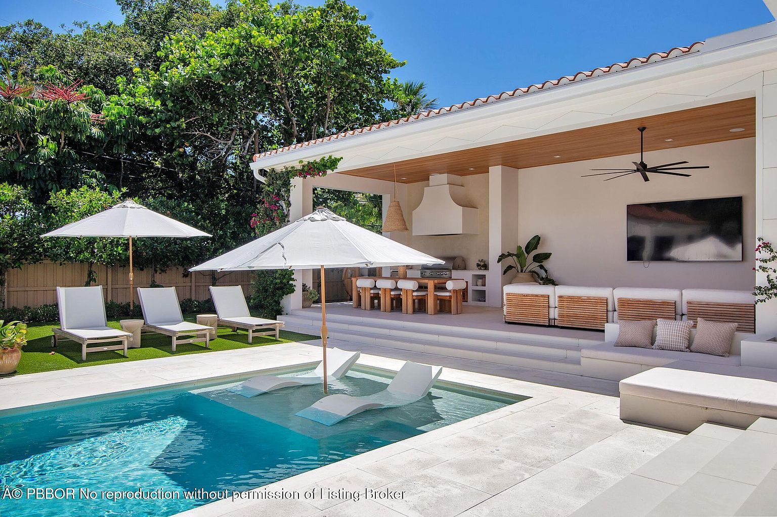 This image showcases a luxurious outdoor pool and patio area, perfect for relaxation and entertainment. The pool features built-in lounge chairs, and the patio includes comfortable seating, an outdoor kitchen, and a covered dining area. Lush landscaping surrounds the space, creating a private and serene atmosphere.