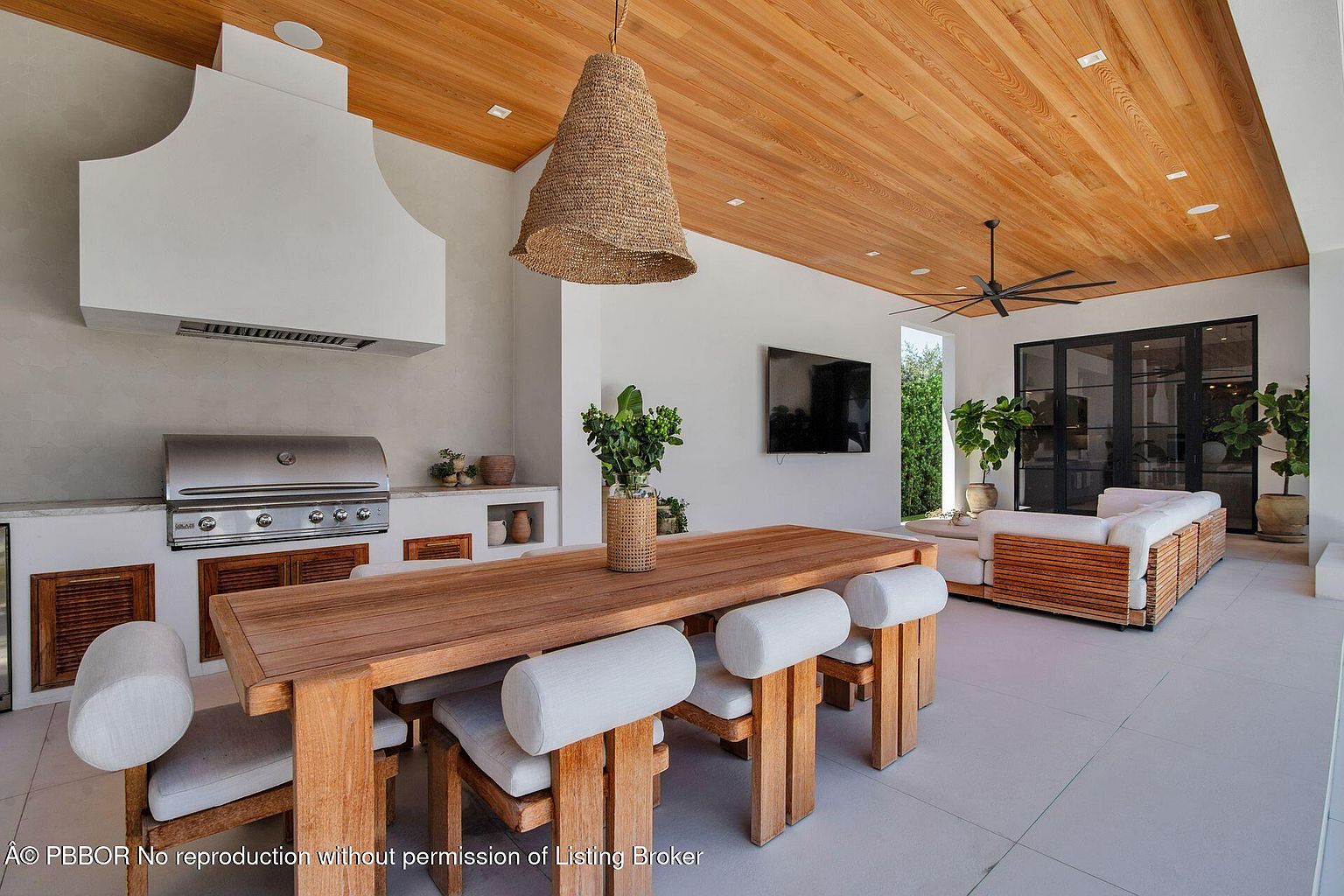 This image showcases a luxurious outdoor patio area, featuring a built-in kitchen with a stainless steel grill and custom cabinetry. A large wooden dining table with cushioned chairs sits under a woven pendant light, creating an inviting space for outdoor dining. The area also includes a comfortable seating area with a wooden frame sofa and a large screen television, perfect for entertaining.