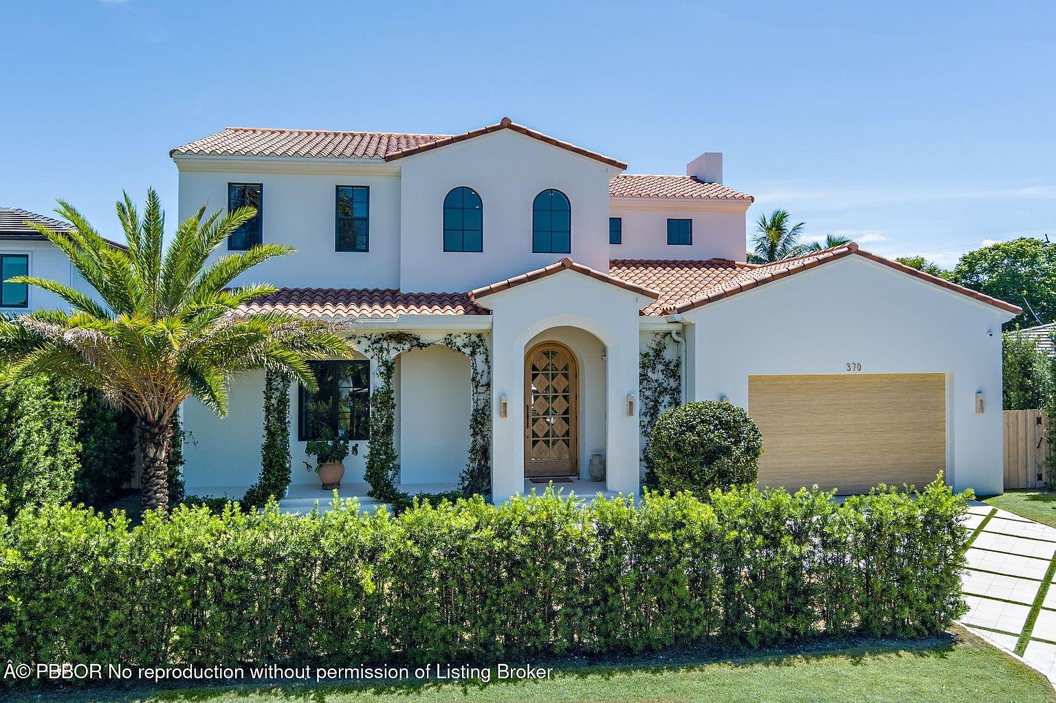 This is a front exterior view of a two-story Mediterranean-style house. The house features a white facade, a red tile roof, arched windows, and a prominent front door with decorative details. Lush green landscaping and a manicured lawn enhance the property's curb appeal, creating an inviting and luxurious impression.