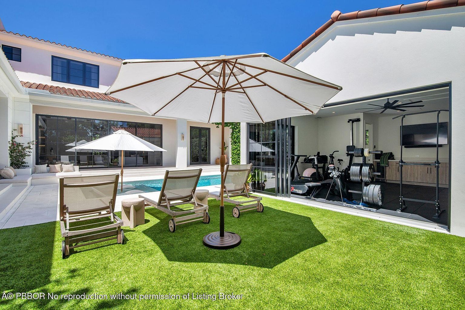 This image showcases a beautifully landscaped backyard featuring a well-manicured lawn, a swimming pool, and lounge chairs under a large umbrella. The property includes a modern home with large glass doors leading to an indoor gym, creating a seamless indoor-outdoor living experience. The perspective is from ground level, emphasizing the depth and breadth of the yard.