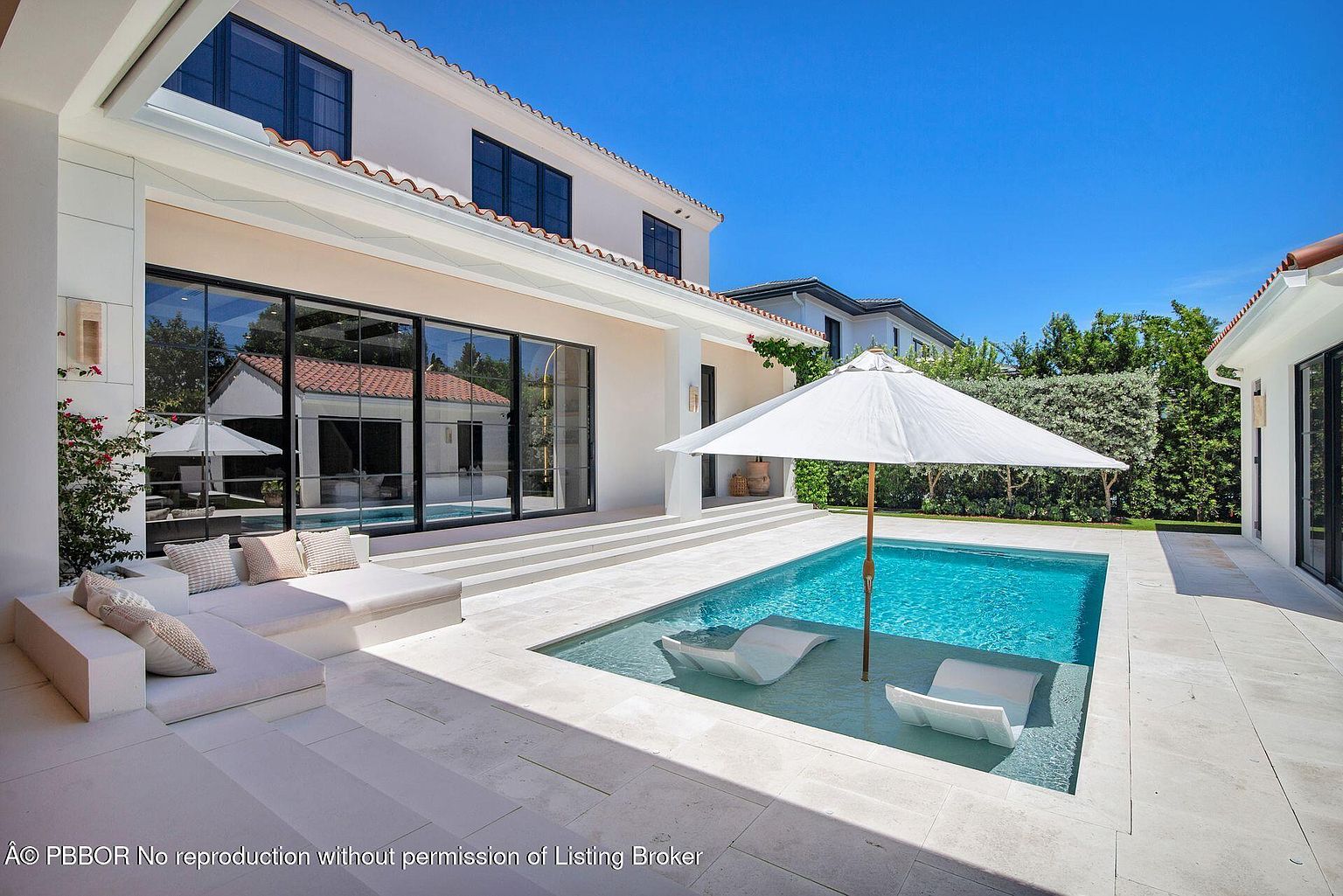This image showcases a luxurious backyard pool area. The pool features built-in lounge chairs and is shaded by a large white umbrella. The surrounding patio is made of light-colored stone, and a modern house with large glass doors and windows overlooks the pool area, creating a serene and upscale outdoor living space.
