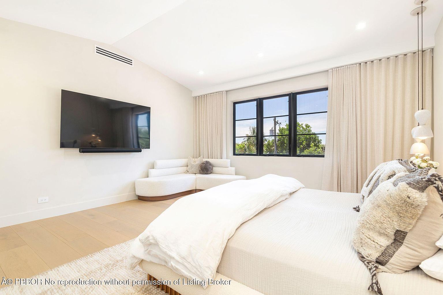 This primary bedroom features a modern aesthetic with light wood flooring and neutral tones. A large window provides natural light, complemented by sheer curtains. The room includes a wall-mounted TV, a unique curved sofa, and a bed with decorative pillows, creating a comfortable and stylish retreat.