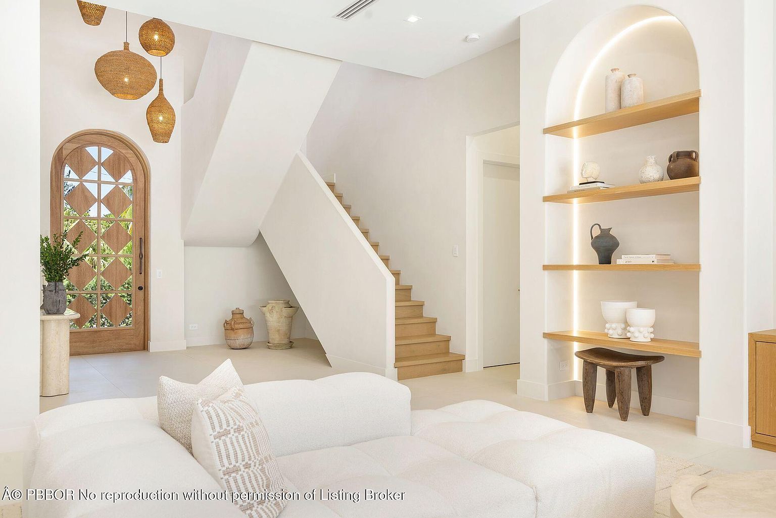 This interior shot showcases a bright and airy living room with a focus on natural light and neutral tones. The room features a comfortable white sectional sofa, a decorative arched doorway, and a staircase leading to the upper level. A built-in shelving unit with decorative items adds visual interest, while unique pendant lights hang above the entrance, creating a welcoming and stylish atmosphere.