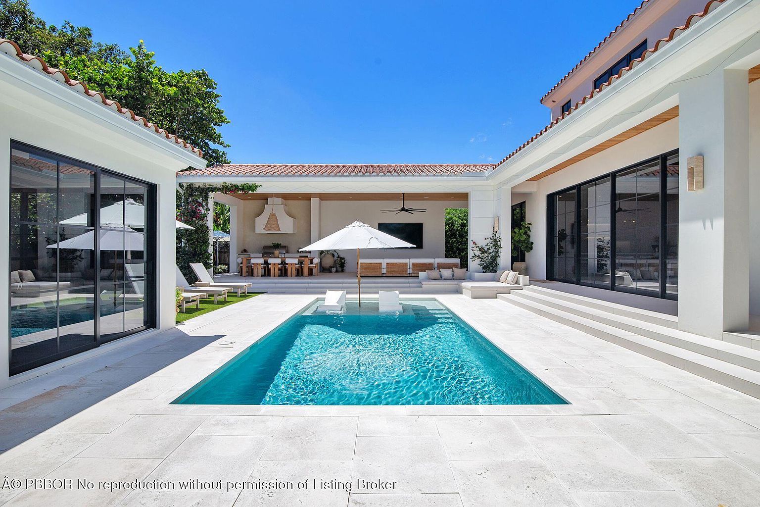 This image showcases a luxurious backyard featuring a rectangular pool with crystal clear water, surrounded by light-colored stone tiling. The pool area is flanked by modern white structures with black-framed glass doors, leading to indoor spaces. An outdoor living area with a dining table and lounge seating is visible under a covered patio, creating an inviting space for relaxation and entertainment.