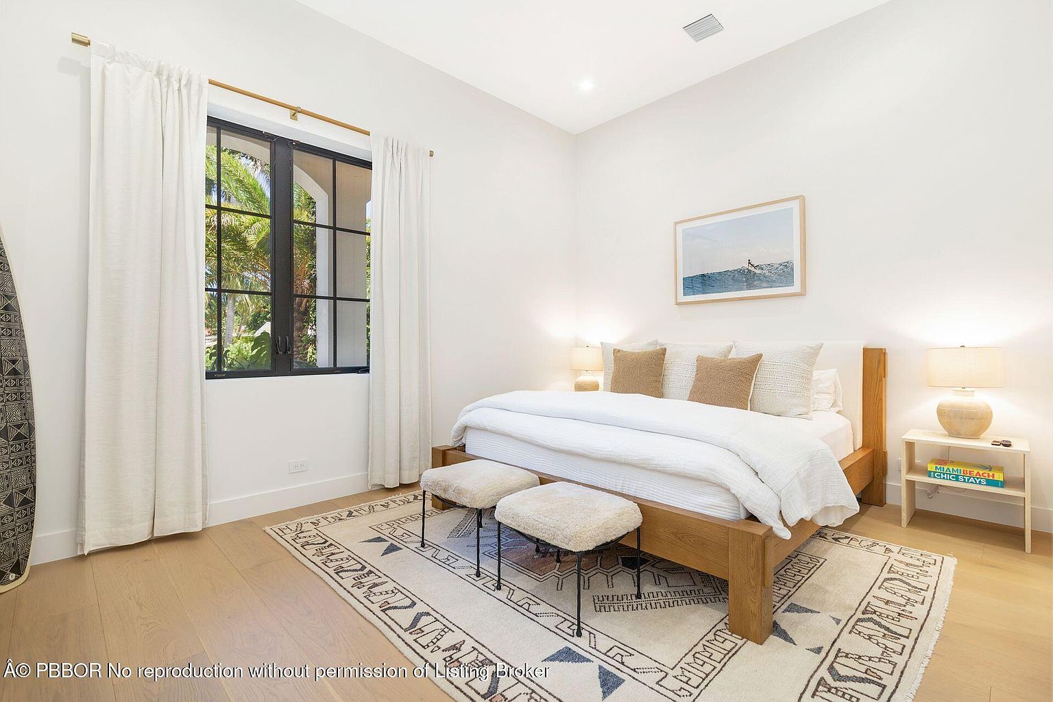 This is a bright and airy primary bedroom featuring a wooden bed frame with white linens and decorative pillows. The room is well-lit by natural light from a large window with white curtains, complemented by bedside lamps on a small side table. A patterned rug adds texture to the wooden floor, and two small stools are placed at the foot of the bed, creating a cozy and inviting atmosphere.