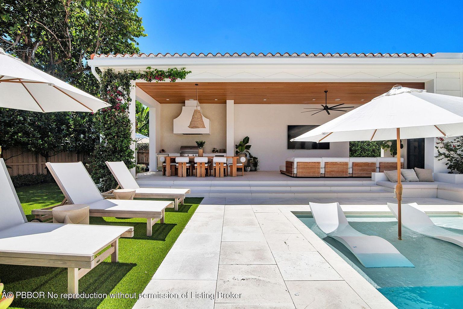 This image showcases a luxurious outdoor pool and patio area, featuring modern lounge chairs both on the patio and in the water. An outdoor kitchen and dining area are visible under a covered structure, complete with a grill, dining table, and seating. The space is designed for relaxation and entertainment, with a seamless blend of indoor and outdoor living.