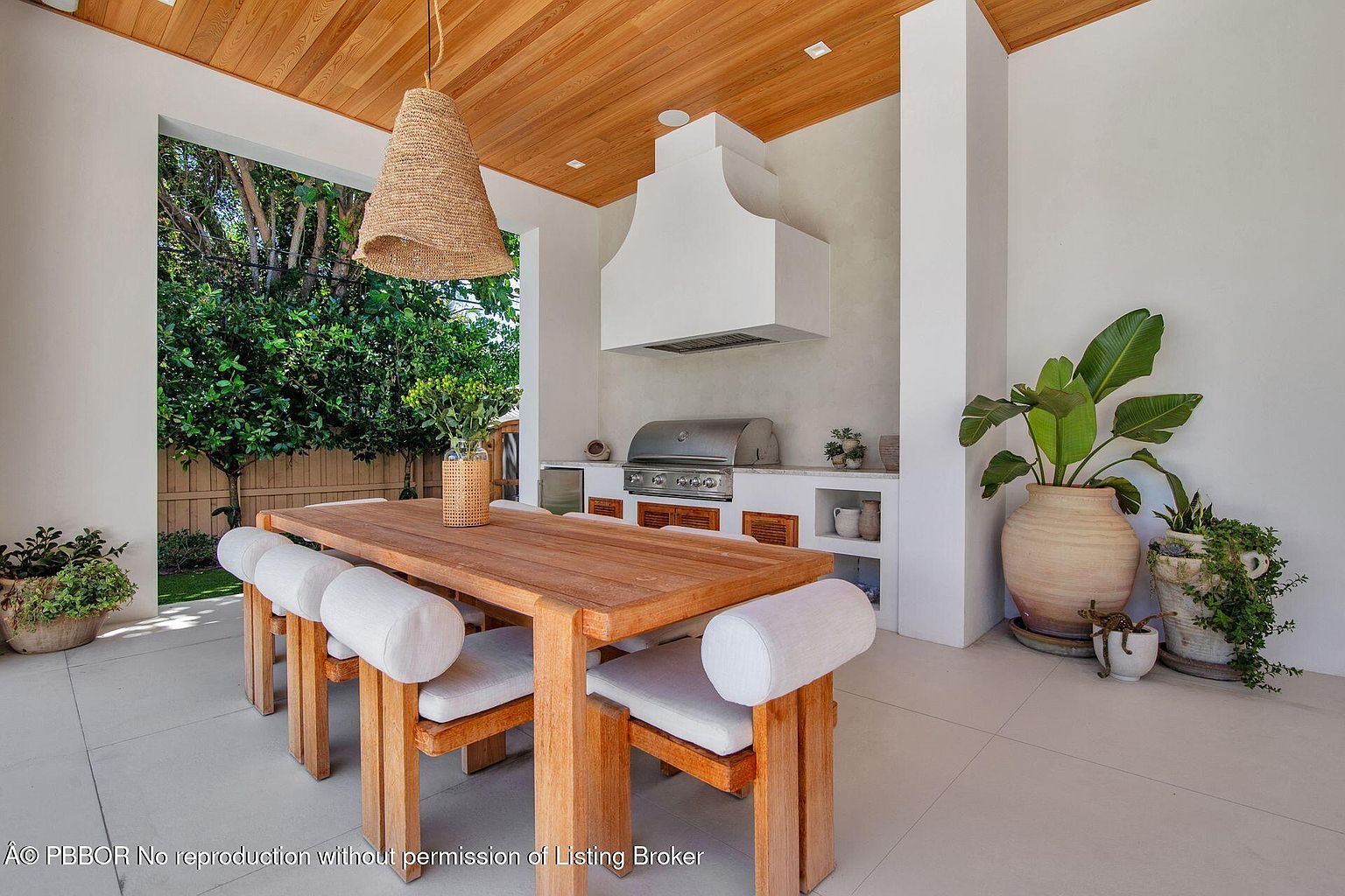 This image showcases an outdoor dining and kitchen area under a covered patio. The space features a wooden dining table with cushioned seating, a built-in outdoor kitchen with a stainless steel grill and hood, and decorative plants in large pots. The design is modern and inviting, perfect for outdoor entertaining.