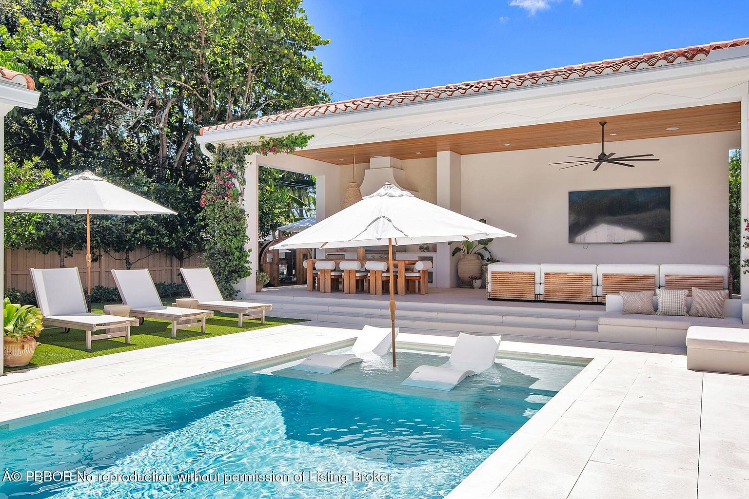 This image showcases a luxurious outdoor pool and patio area. The pool features built-in lounge chairs, and the patio includes a covered outdoor living space with a dining area, a seating area with a large television, and a ceiling fan. The design is modern and upscale, creating an inviting space for relaxation and entertainment.