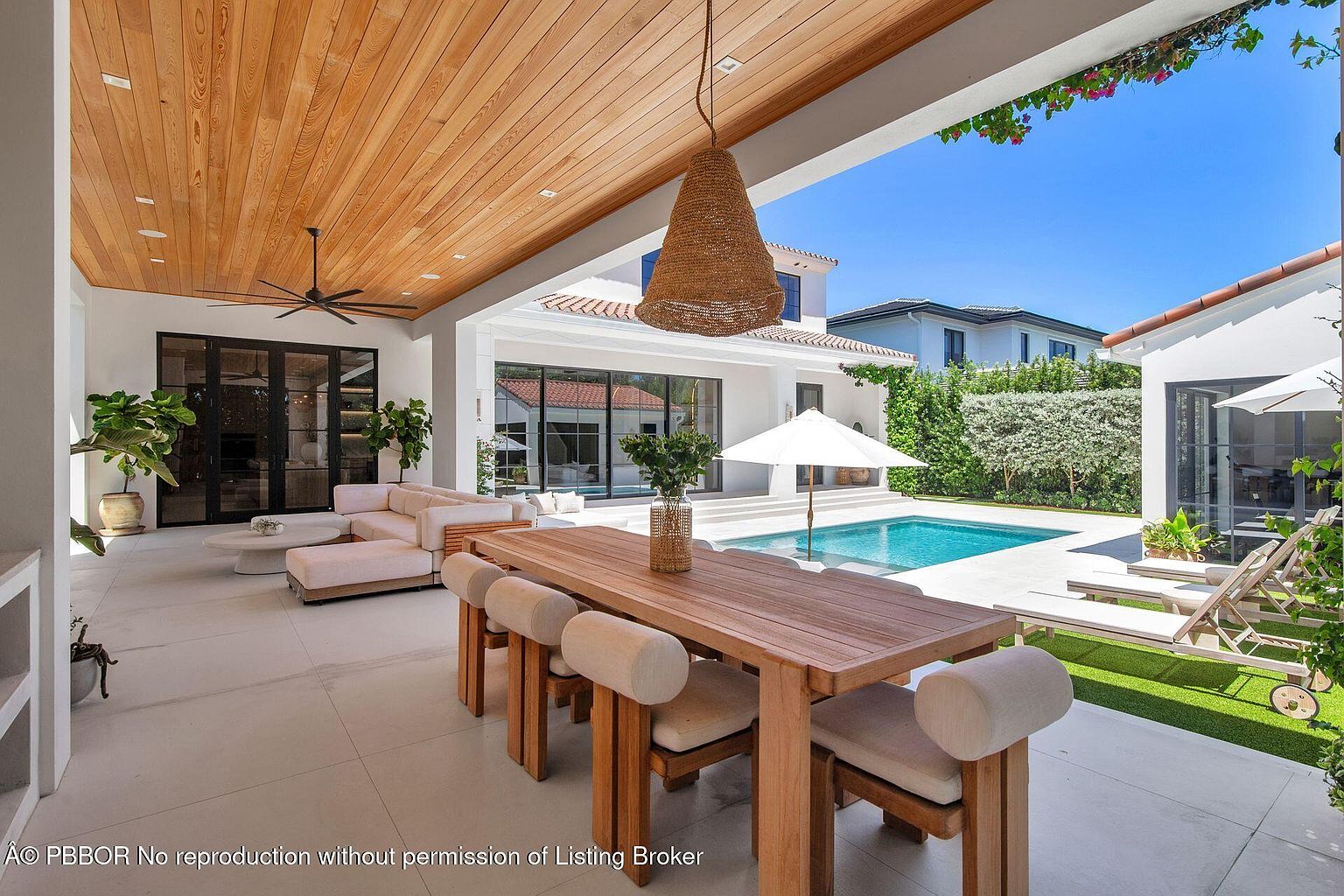 This image showcases a luxurious outdoor patio area, featuring a wooden dining table with cushioned chairs, a comfortable lounge area with a sectional sofa, and a view of a swimming pool. The patio is covered by a wooden ceiling with a unique pendant light, creating a shaded and elegant space for relaxation and entertainment. The backyard includes a pool, lounge chairs, and manicured greenery, enhancing the property's appeal.