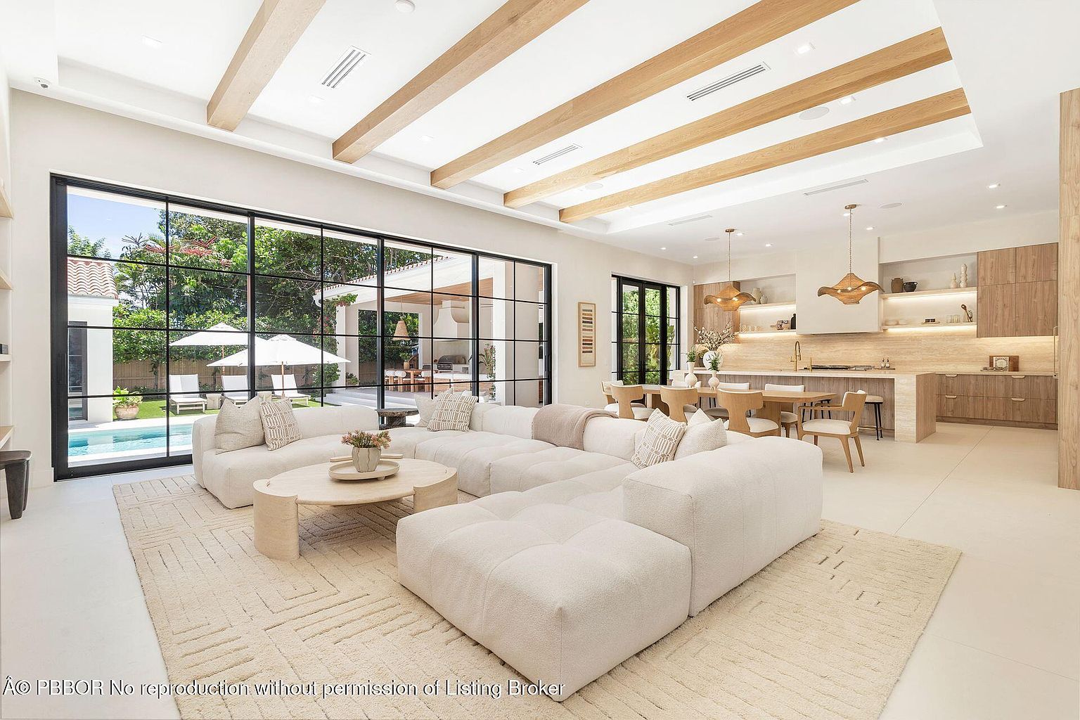 This is an interior shot of a modern living room that seamlessly blends with the kitchen and dining area. The living room features a large, modular white sofa, a round coffee table, and a light-colored rug. Large black-framed windows offer a view of the outdoor pool and patio area, while exposed wooden beams add architectural interest to the ceiling. The kitchen area includes wooden cabinetry and modern pendant lighting, creating a cohesive and stylish living space.