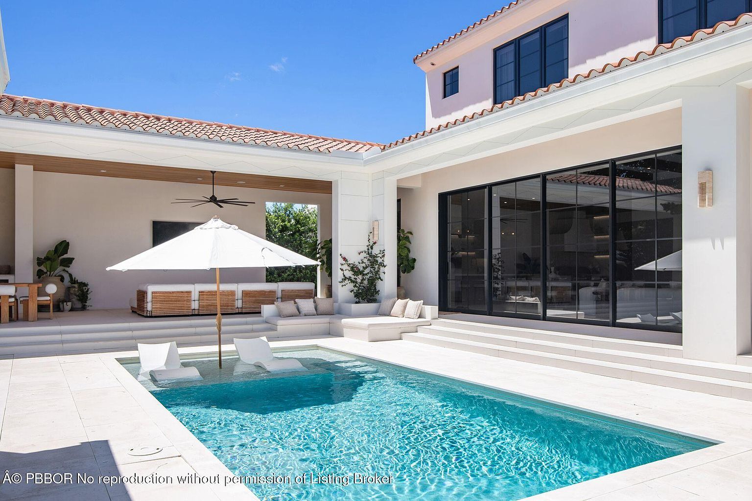 This image showcases a luxurious outdoor pool area, featuring a rectangular pool with clear blue water and built-in lounge chairs. The surrounding patio is paved with light-colored tiles, and a white umbrella provides shade. The architecture includes a covered outdoor living space with comfortable seating and a modern home with large glass doors, creating a seamless indoor-outdoor transition.