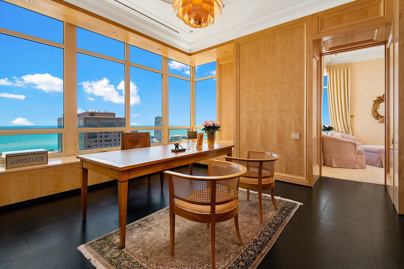 This is an interior shot of a luxurious office or study. The room features a large wooden desk, two cane-backed chairs, and a large window with a view of the water and city skyline. The walls are paneled in a light wood, and the floor is a dark hardwood, creating a sophisticated and elegant atmosphere.