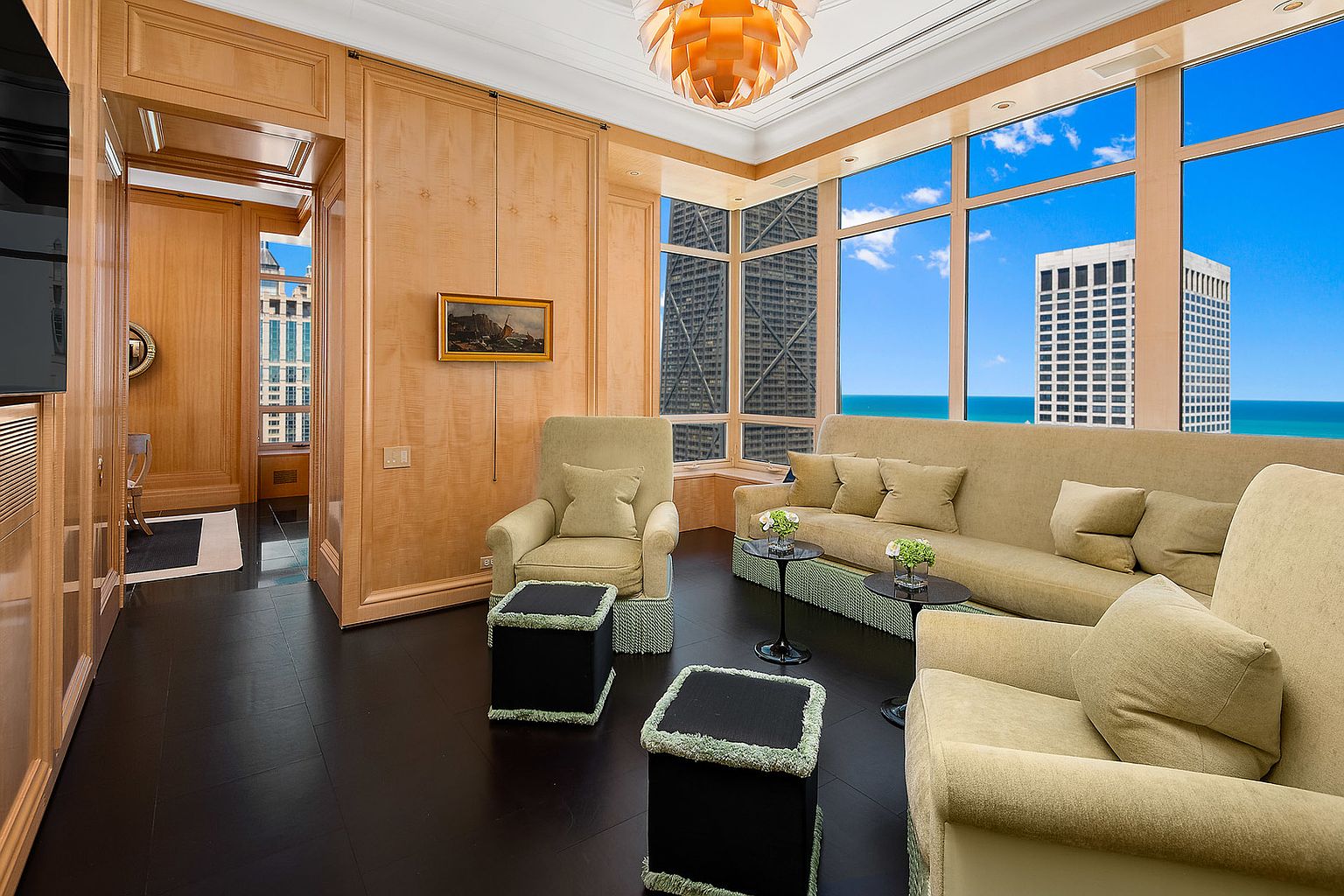 This interior shot showcases a luxurious living room with dark wood flooring, light wood paneled walls, and large windows offering city and water views. The room is furnished with comfortable seating, including armchairs and a sofa, complemented by stylish ottomans and side tables. A unique light fixture hangs from the ceiling, adding a touch of modern elegance.