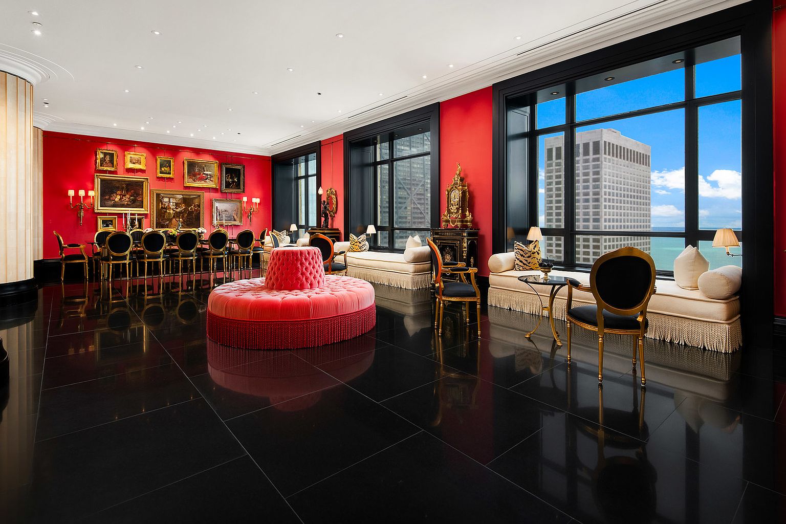 This opulent living room features a striking red accent wall adorned with classic paintings, complemented by black window frames offering city views. The room is furnished with a large, round red ottoman, elegant seating arrangements, and a long dining table with gold-trimmed chairs, all set against a backdrop of polished black tile flooring. The overall impression is one of luxury and sophistication.