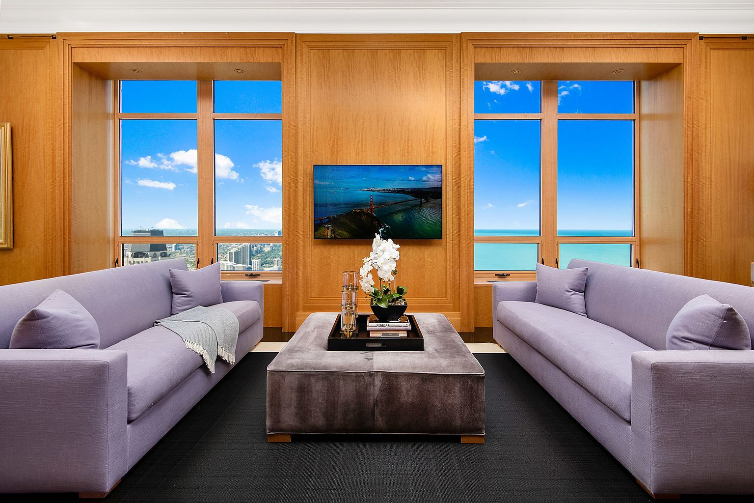 This is an interior shot of a luxurious living room featuring two matching lavender sofas facing each other, separated by a large, plush ottoman. The room is framed by wood-paneled walls with large windows offering a stunning view of the city and ocean. A flat-screen television is mounted on the central wood panel, adding a modern touch to the classic design.