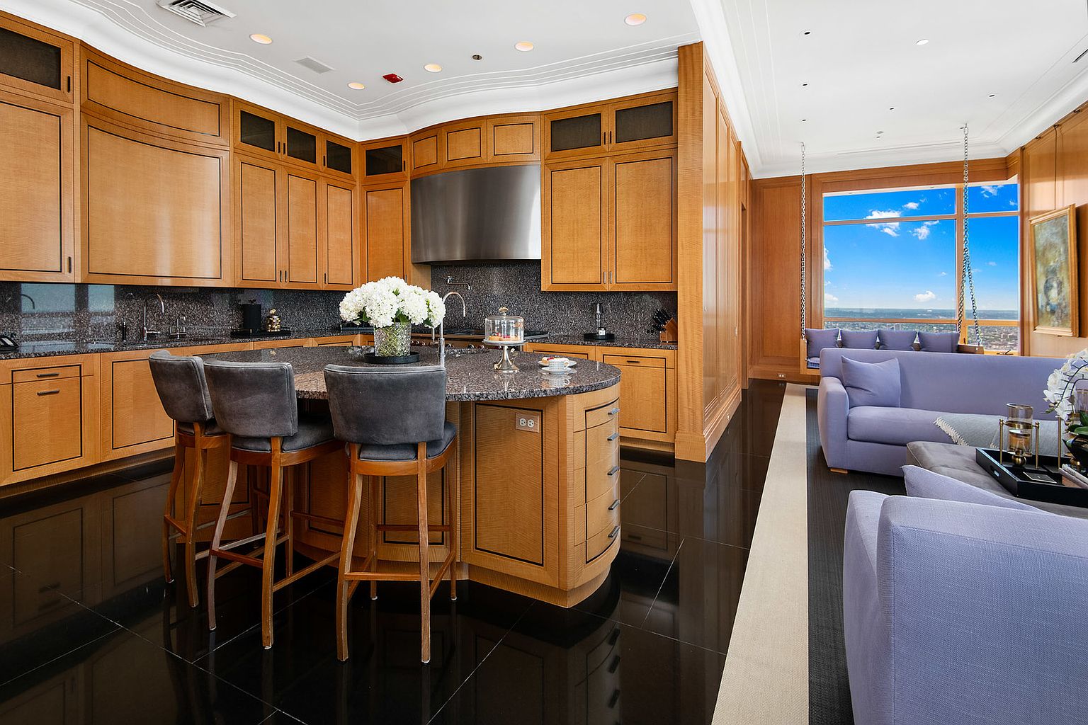 This interior shot showcases a luxurious kitchen that seamlessly blends into a living area. The kitchen features custom wood cabinetry, granite countertops, and a stainless steel range hood. The living area includes plush seating and a large window with a city view, creating an open and inviting space.