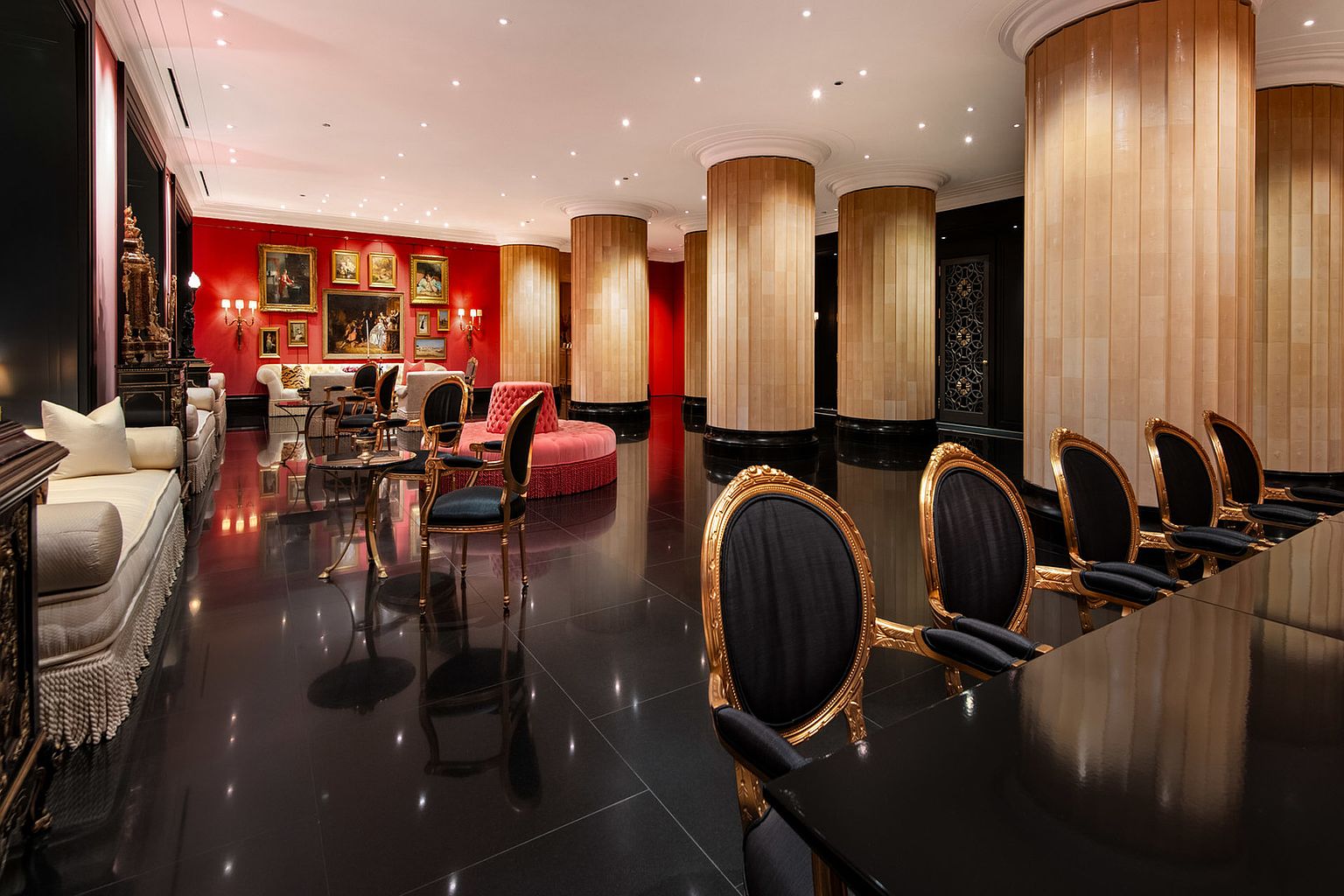 This opulent living room features a striking red accent wall adorned with classic paintings, complemented by elegant seating arrangements including plush sofas and ornate chairs with gold frames. The room is characterized by its high-gloss black tile flooring, which reflects the light from the recessed ceiling lights and wall sconces, creating a luxurious and dramatic ambiance. Large cylindrical columns add architectural interest, and the overall impression is one of sophisticated grandeur.